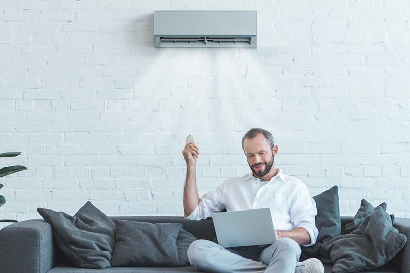 Man sitting on couch turning on ductless ac unit with remote controller.